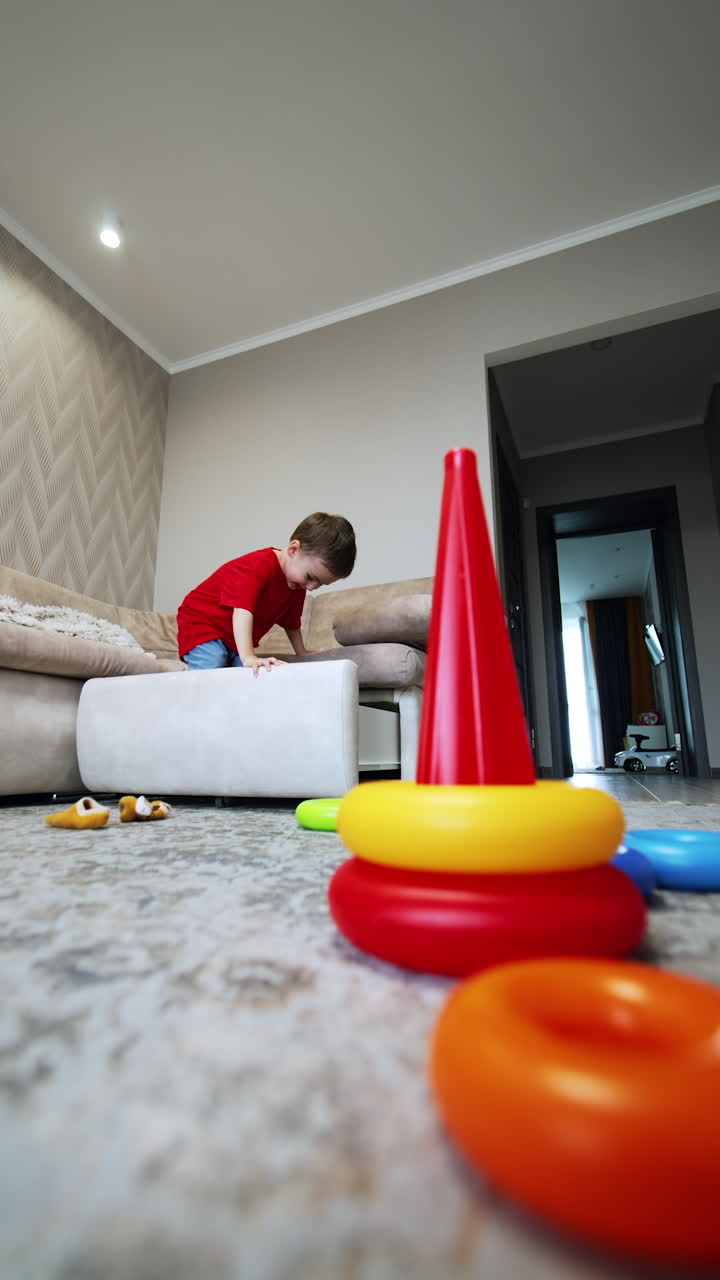 Lovely Caucasian baby boy in red t-shirt and blue pants opens a chest in the sofa and climbs inside it. Cute toddler having fun playing at home. Vertical video.