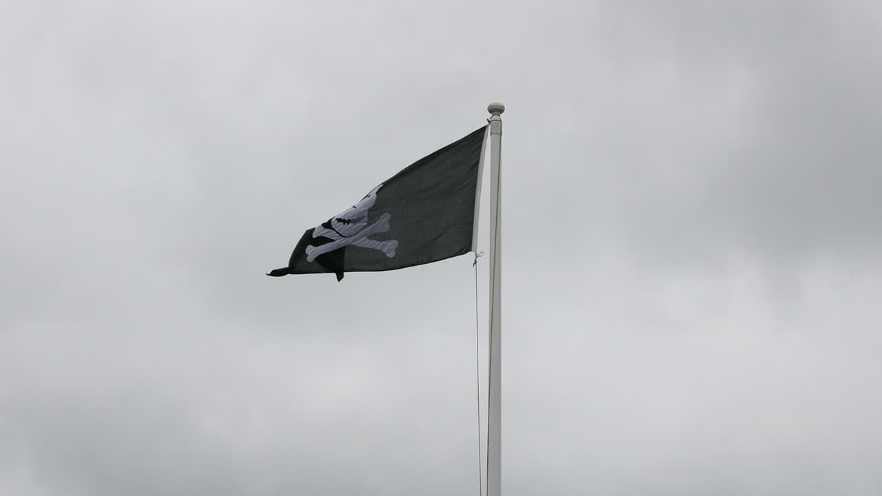calavera de pirata y bandera de huesos cruzados volando en el viento en un poste de bandera