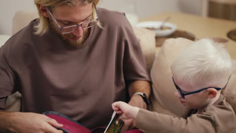 Close-up-shot-of-a-blond-man-with-a-beard-and-glasses-helping-his-little-albino-son-with-white-hair-in-blue-glasses-pack-his-backpack-before-going-to-school-in-the-morning