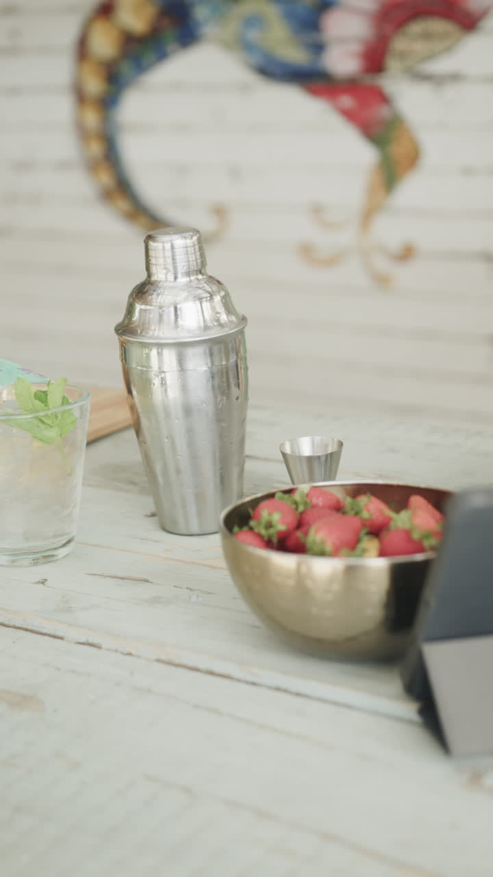 Vertical video of tablet, cocktail shaker, drinks and ingredients on bar at beach bar, slow motion