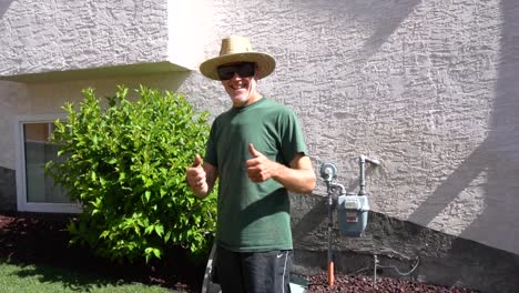 Landscaper-with-sunglasses,-mask,-and-straw-hat-give-two-thumbs-up-after-completing-a-job