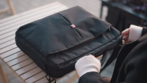 close-up rear view of a lady with ring-adorned fingers and polished nails unzipping a black bag, revealing a laptop inside, set on a wooden table in an outdoor setting