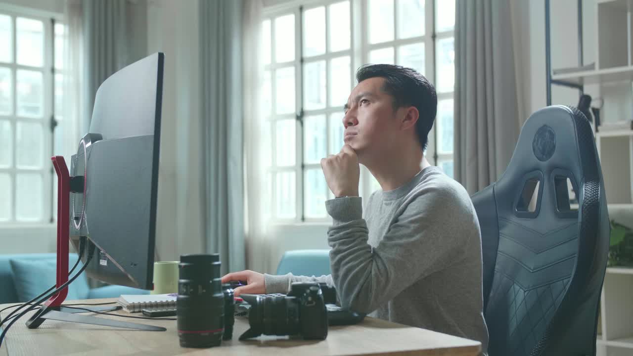 Asian Cameraman In Long Sleeved T-Shirt Thinking While Using Desktop Computer For Working At Home.