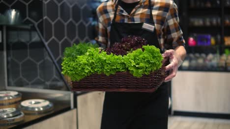 seller man in apron in supermarket walking by vegetables aisle with box of fresh greens to arrange. caucasian worker in local supermarket holding box of greens. close up. slow motion