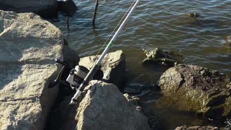 Fishing-rod-set-up-waiting-for-fish-to-bite-bait,-rocky-lake-coastline