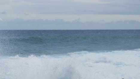 wind-stirred swell rolls toward a rocky shoreline, white water heaving and spreading under gray, overcast skies, conveying coastal motion, moody weather, and the steady pulse of the open sea