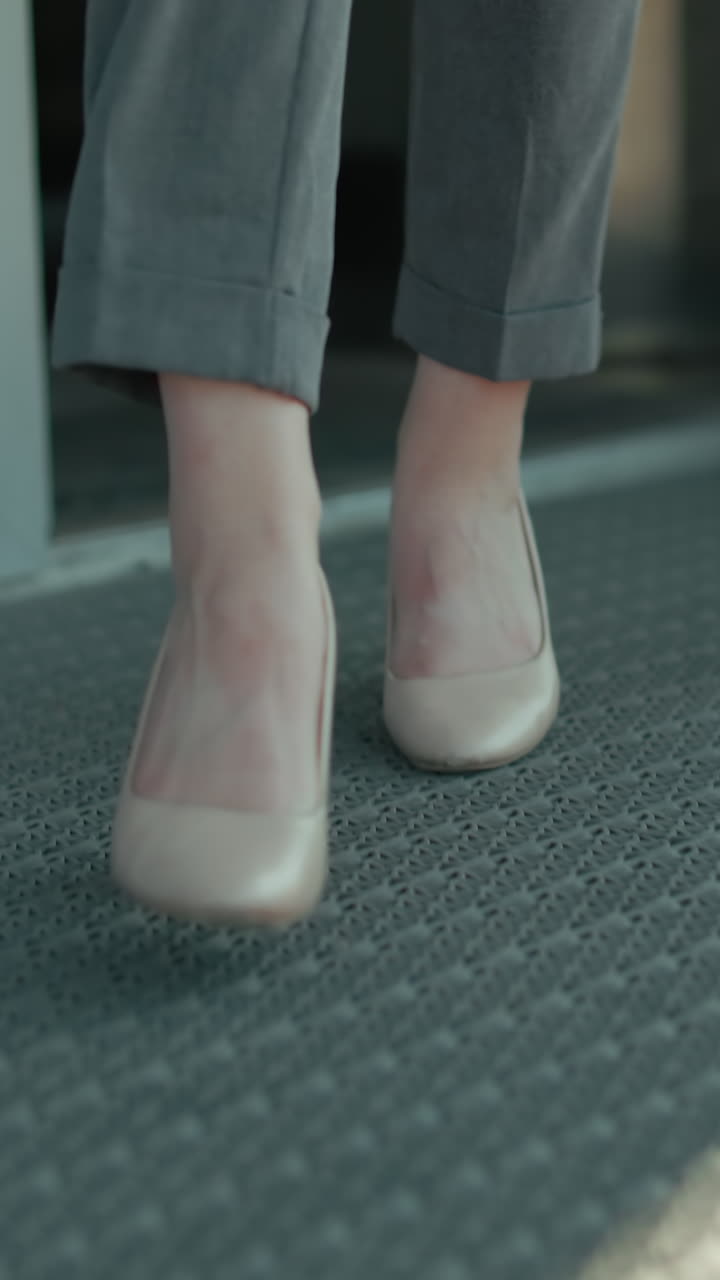 Lower angle view of professional woman in heels stepping out of building through glass door, focus on movement, footwear, modern office environment