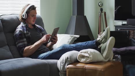 happy caucasian man sitting on sofa in living room wearing headphones and using smartphone