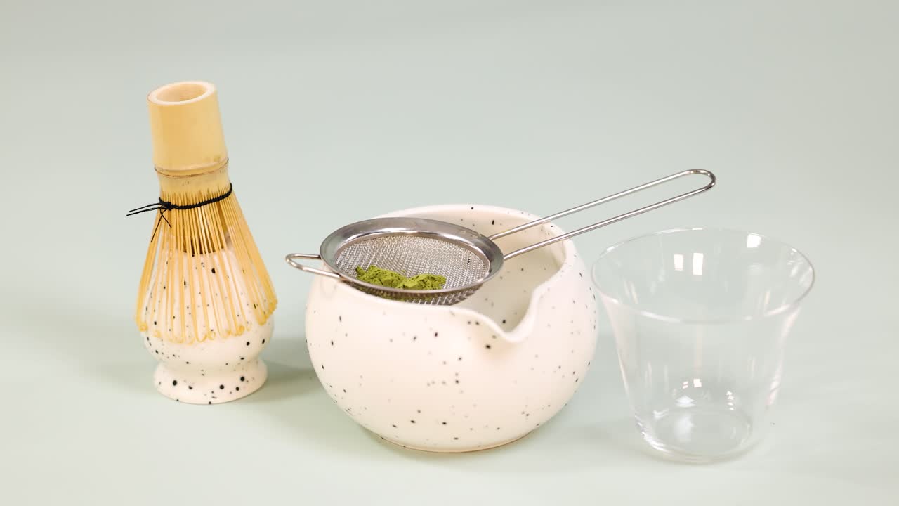 Hand sifts matcha powder into bowl using bamboo scoop, with traditional whisk and soft lighting