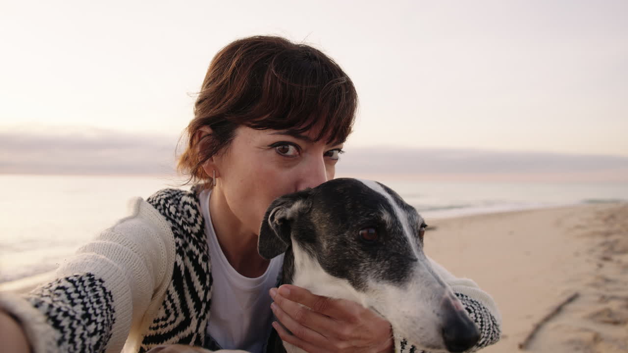 Woman taking a selfie with her dog on the beach at sunset. Close up shot with warm light and direct eye contact with camera. Lifestyle and pet companionship
