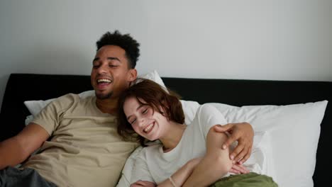 A-happy-young-girl-with-a-bob-hairstyle-in-a-white-T-shirt-lies-on-the-bed-with-her-boyfriend-with-Black-skin-color-and-watches-a-comedy-on-a-gray-laptop-in-the-morning