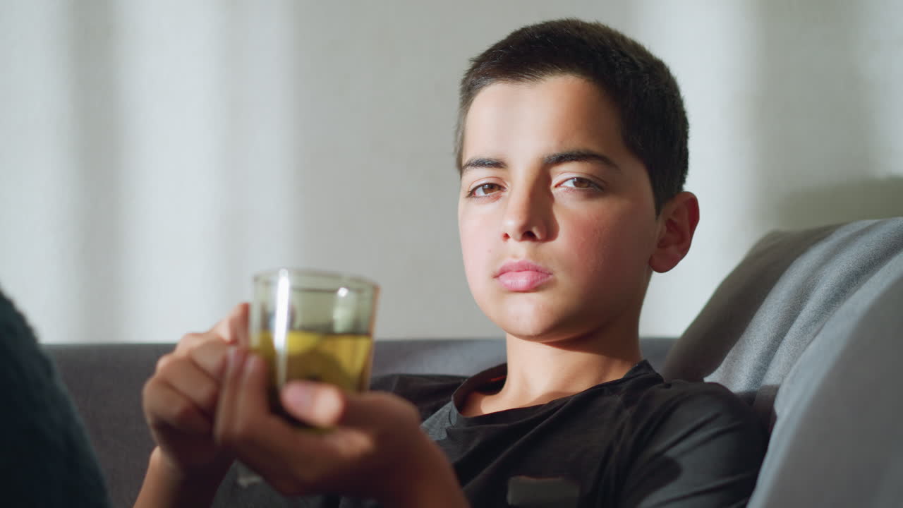 Boy sitting on a couch holding tea cup close to mouth, staring at the camera with a tired, sickly expression, conveying a sense of illness or discomfort while resting