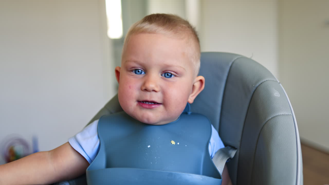 Adorable blond boy with silicone utensils sits on a high chair for feeding. Joyful child drinks deliciously and smiles. Child development. Children's furniture for feeding