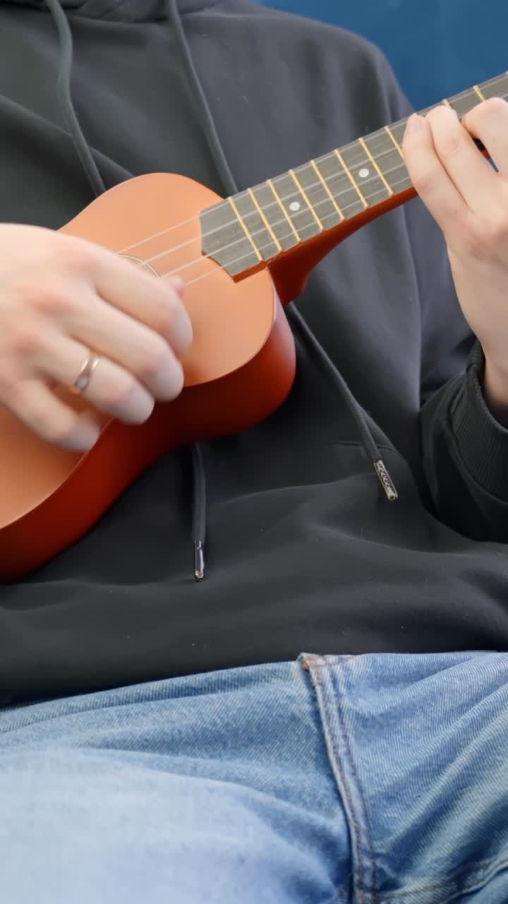 Hand plucks strings of wooden ukulele gently while sitting on chair indoors, vertical