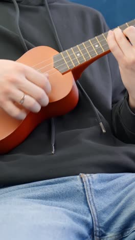 hand plucks strings of wooden ukulele gently while sitting on chair indoors, vertical