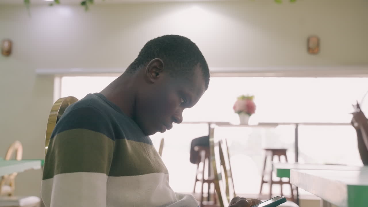 Black man in cafe checking phone, waiting for date nervous glance, tapping thumbs, striped sweater, sunlight through window, vase on table, soft backlit interior, quiet anticipation and hopeful