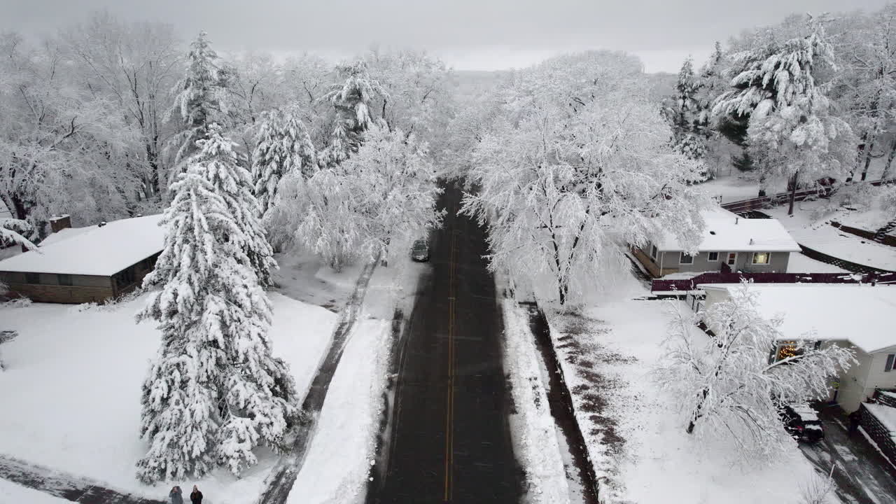 Aerial drone view of two vehicles navigating a snow-covered road during a winter storm with headlights on, showcasing hazardous driving conditions and a serene frozen landscape