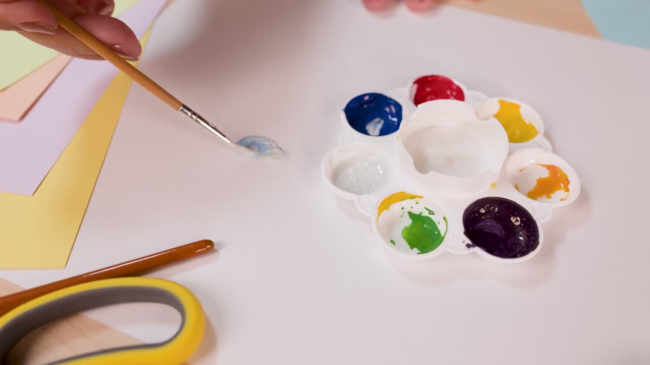 Artist hand mixes blue watercolor paint on palette, bright workspace, top-down view, soft lighting