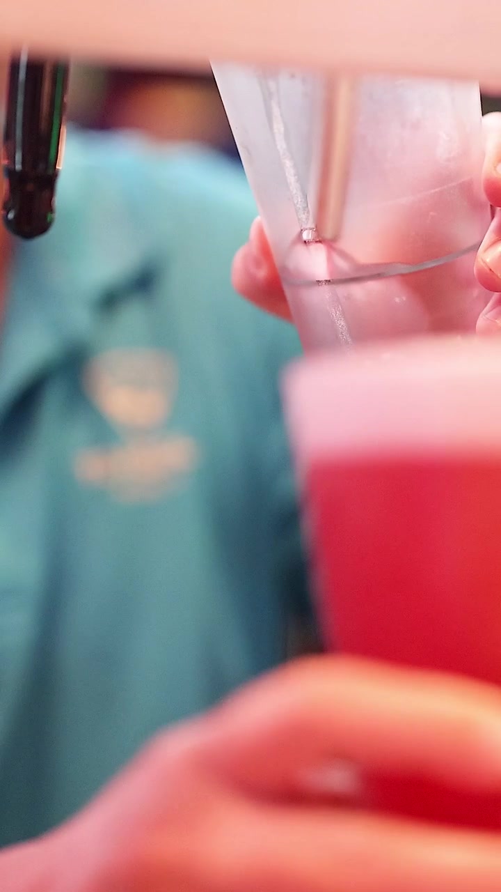 A close-up vertical view of a bartender pouring a vibrant red beverage into a glass.