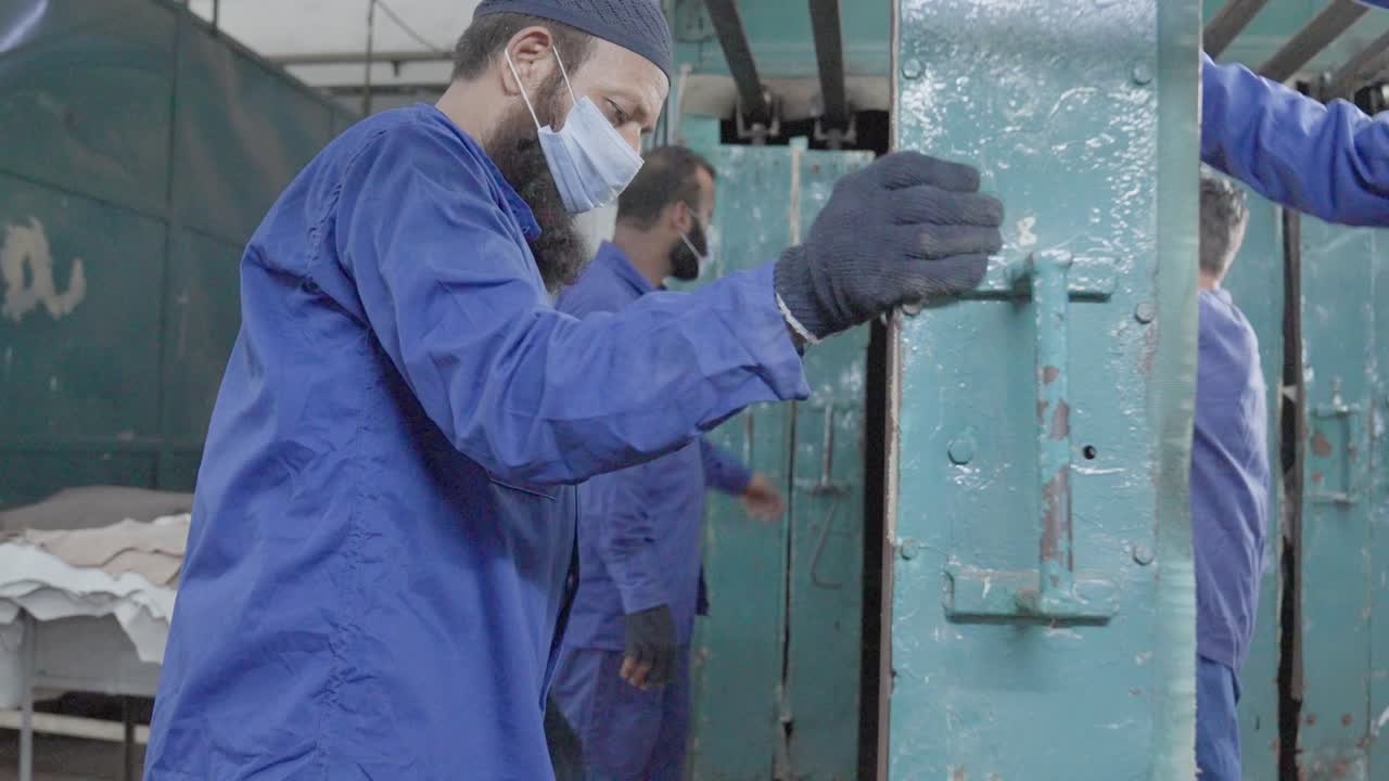 Worker of a leather factory changing metal net to work with skin leather, attached with brackets, static shot, blue clothes, masks and gloves