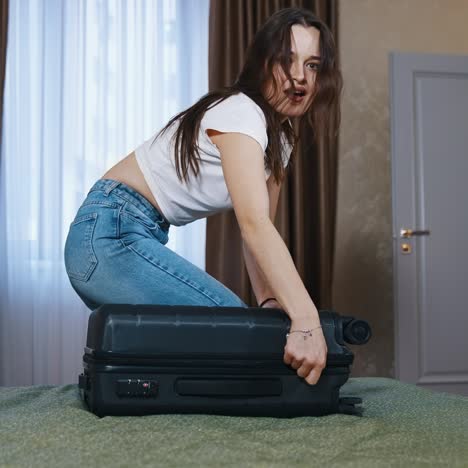 Young woman with travel bag indoors. Pretty girl on bed trying to close the suitcase full of clothes. Female packing luggage in the hotel room