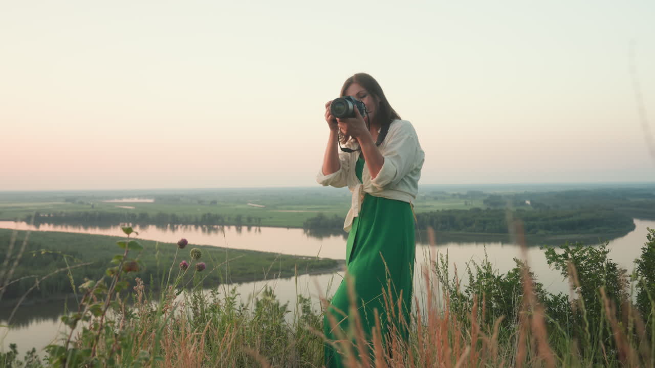 Lady holding camera captures scenic river landscape during golden hour while smiling, dressed in white shirt and green skirt, standing near bushy edge overlooking wide water and distant green terrain