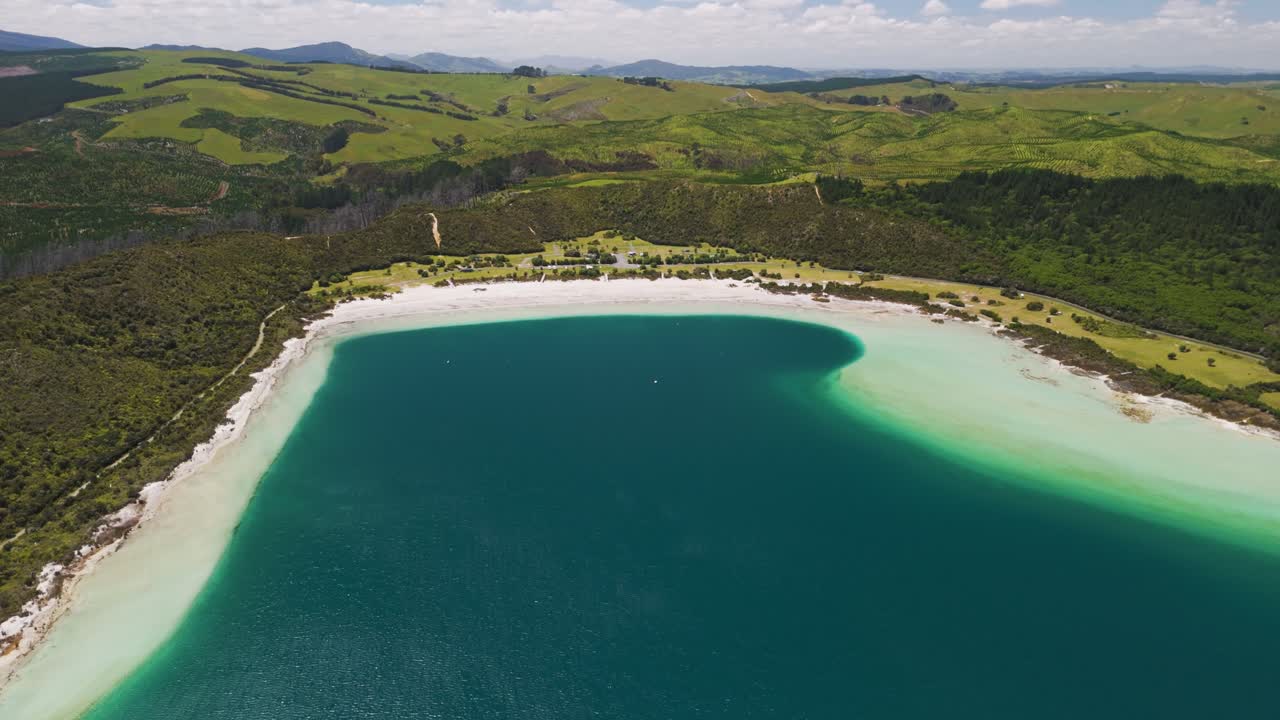 Sweeping orbital view of crescented white sand beach embracing turquoise waters at Kai Iwi Lakes with pastoral hills and distant mountains evoking remote paradise