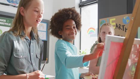 video of diverse, happy schoolchildren standing at easels painting, talking in art class, copy space
