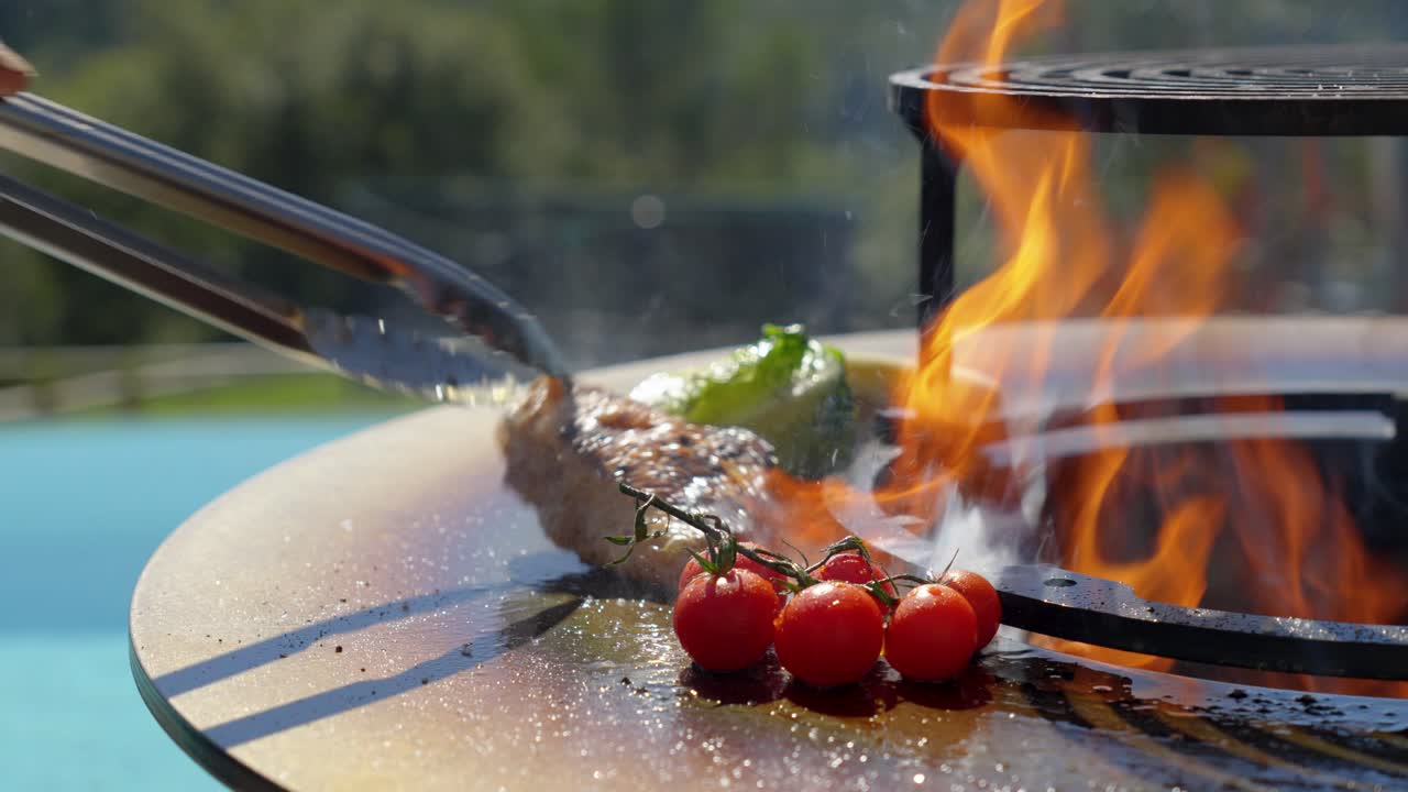 close-up of a brazier with a hand holding tongs and turning food on the hotplate, flame in the centre of the brazier and blurred swimming pool in the background