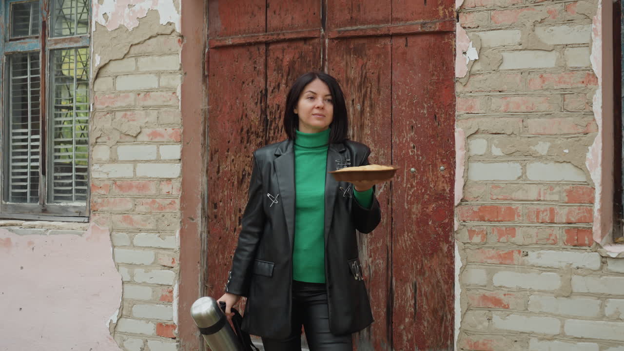 woman in leather jacket holding hat, standing by weathered brick doorway, green sweater visible, vintage camera in hand suggesting traveling photographer, relaxed smile and confident pose, gritty