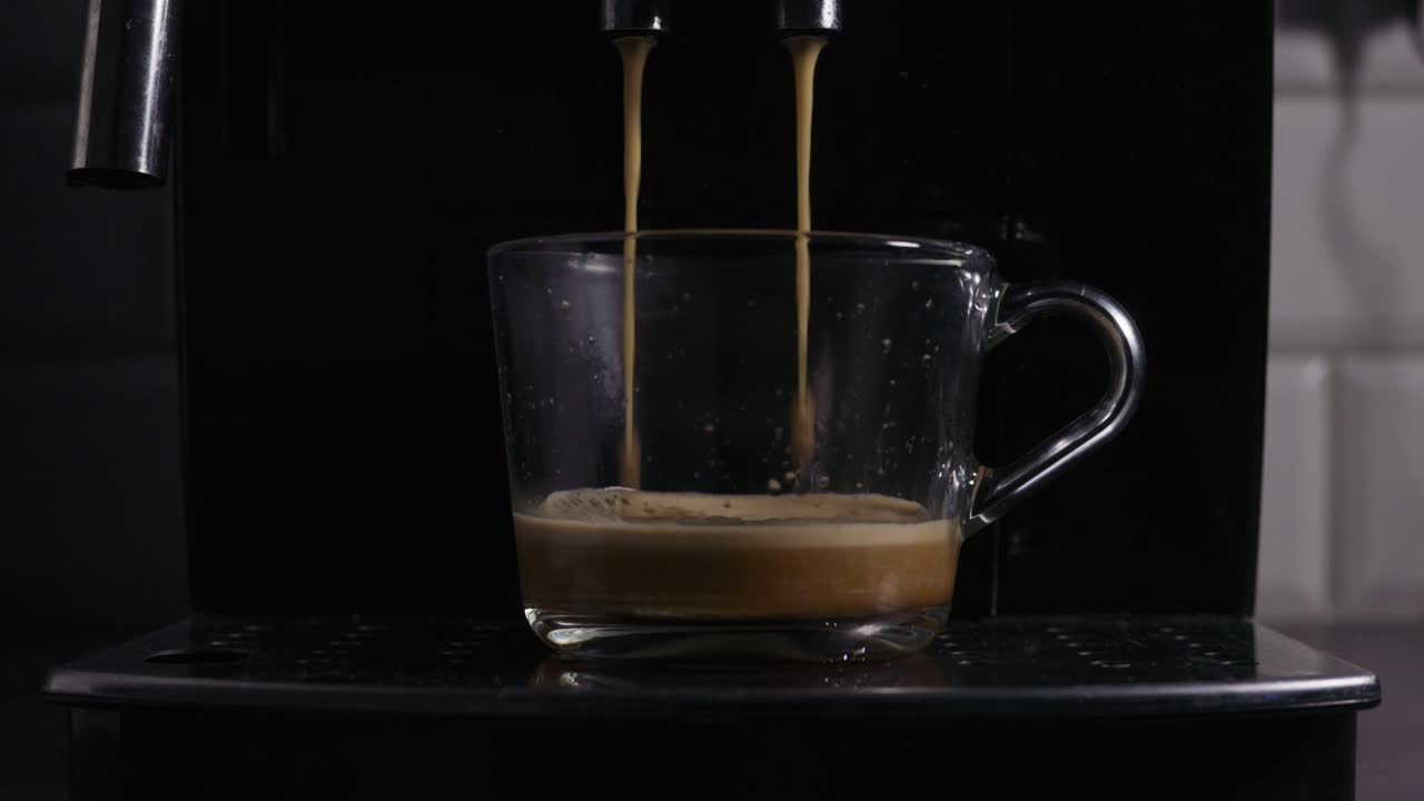 Primo piano di una tazza di vetro che viene riempita con espresso appena fatto da una macchina da caffè. La ricca crema si forma in superficie mentre i doppi getti di caffè si versano delicatamente nella tazza trasparente.