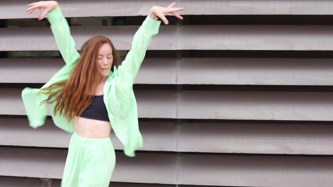 Una mujer con un atuendo verde lima baila alegremente en una ciudad vibrante frente a una pared moderna.