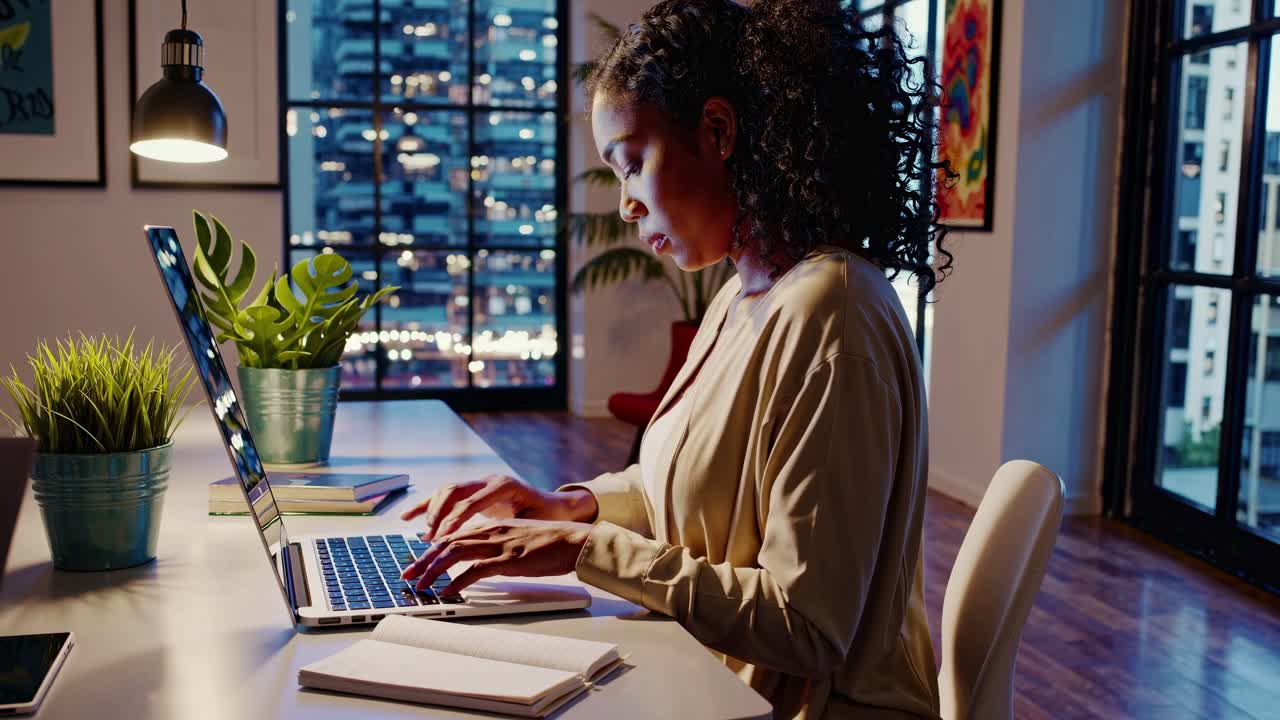 A woman works on a laptop in a modern office at night. Side angle captures city lights. Video style