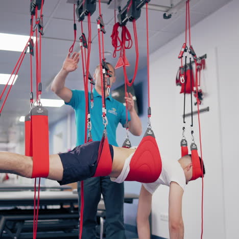 personal trainer controls the process of recovery client on suspension ropes. young man is hanging on special rope system in rehabilitation center. instructor helps his patient to get better.