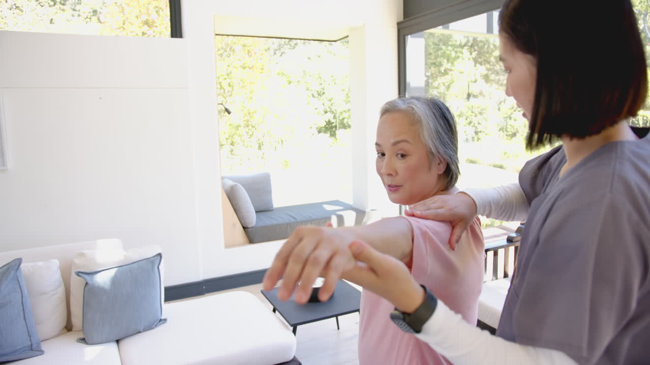 Assisting senior asian woman with shoulder exercises, physiotherapist guiding at home