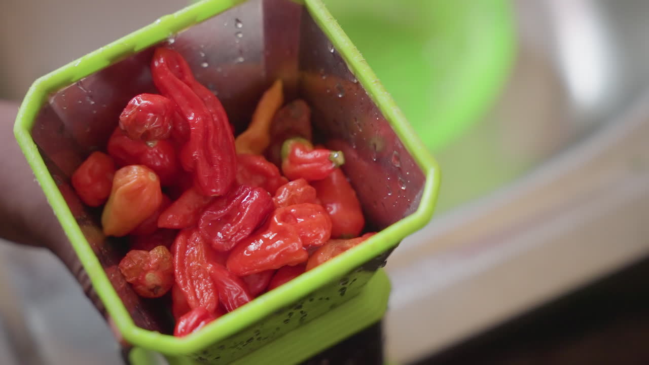 Primo piano di una mano che carica peperoni habanero rugosi in un macinino verde, peperoni rosso vivo, consistenza vivida e pelle lucida, preparazione di calore intenso per salsa in piccole quantità, agitazione e pressione tattile.