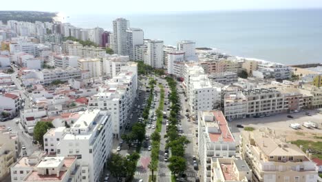 Flying-down-a-beautiful-beach-side-street-lined-with-cars-and-white-hotels-and-luxury-residential-coastal-buildings,-drone-aerial