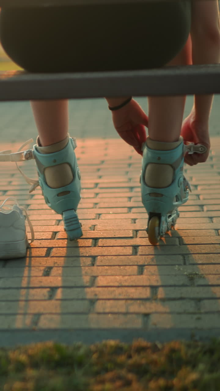 Back view of young lady seated on bench in evening sunlight, removing strap of roller skate with two white sneakers placed beside her on ground
