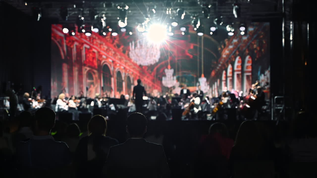 Audience members relax and listen to a symphony orchestra performing in an elegant venue. The stage is decorated with bright lights and a stunning background, creating a magical atmosphere