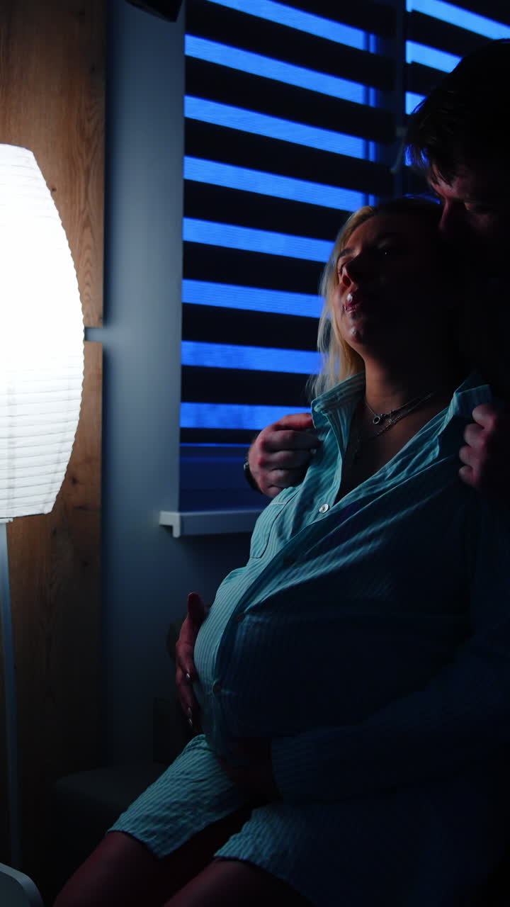 Husband caressing his pregnant wife sitting on the bed. Woman prepares for childbirth in hospital. Dark room backdrop. Vertical video