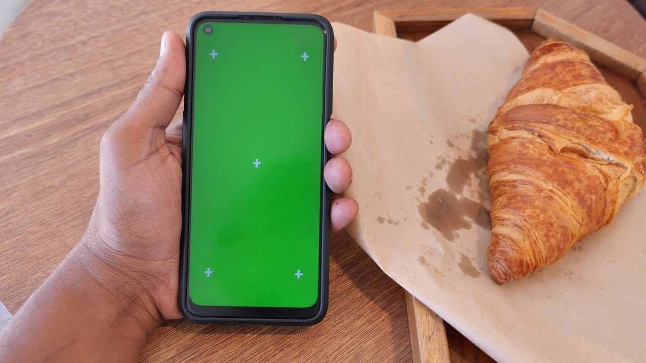 Hand holding a smartphone with green screen next to a croissant