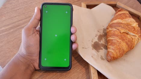 hand holding a smartphone with green screen next to a croissant