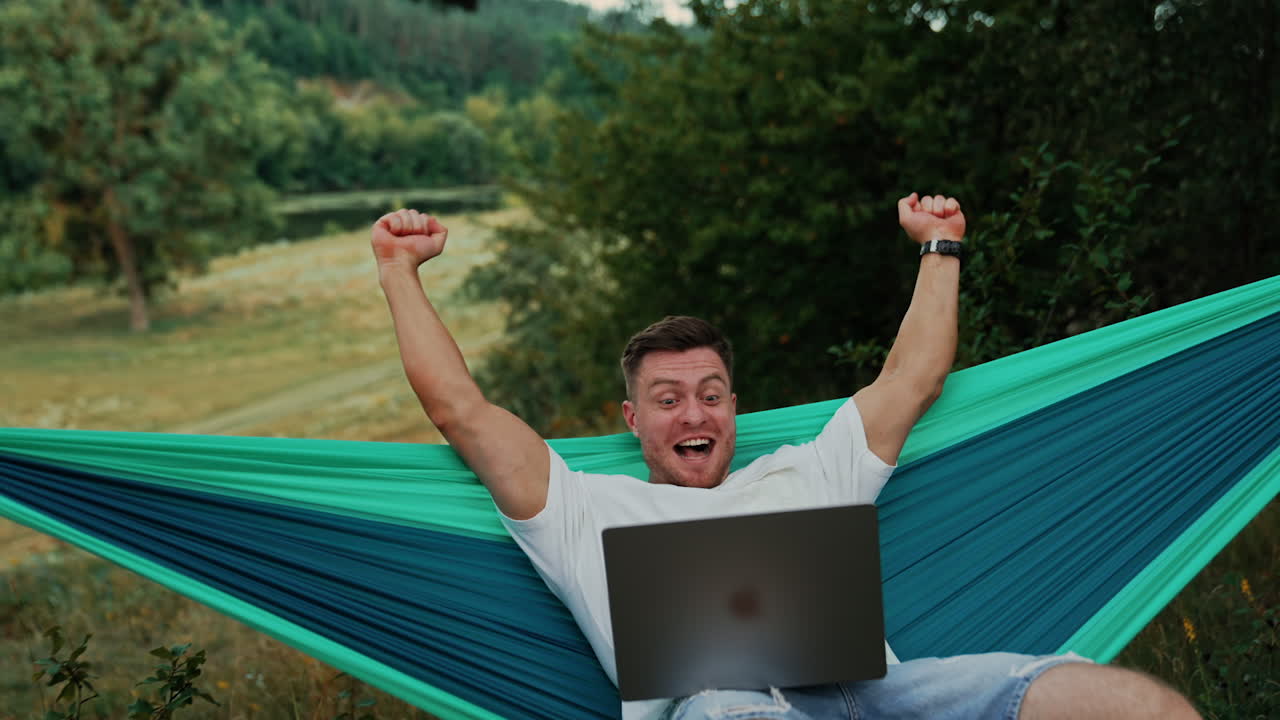 Handsome Caucasian man cheering excitedly working on his laptop. Man sits in a hammock in nature working.