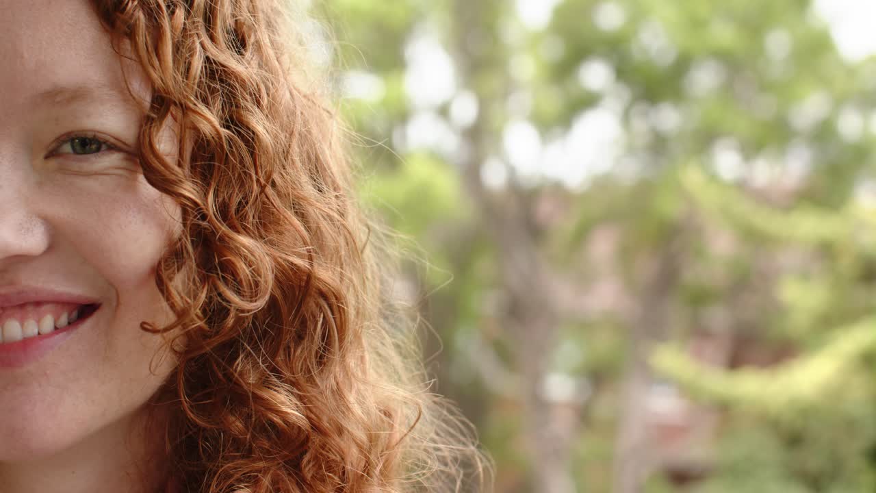 Mujer pelirroja rizada con una sonrisa cada vez más amplia, mostrando los dientes y riendo, apoyada en el jardín, espacio para texto