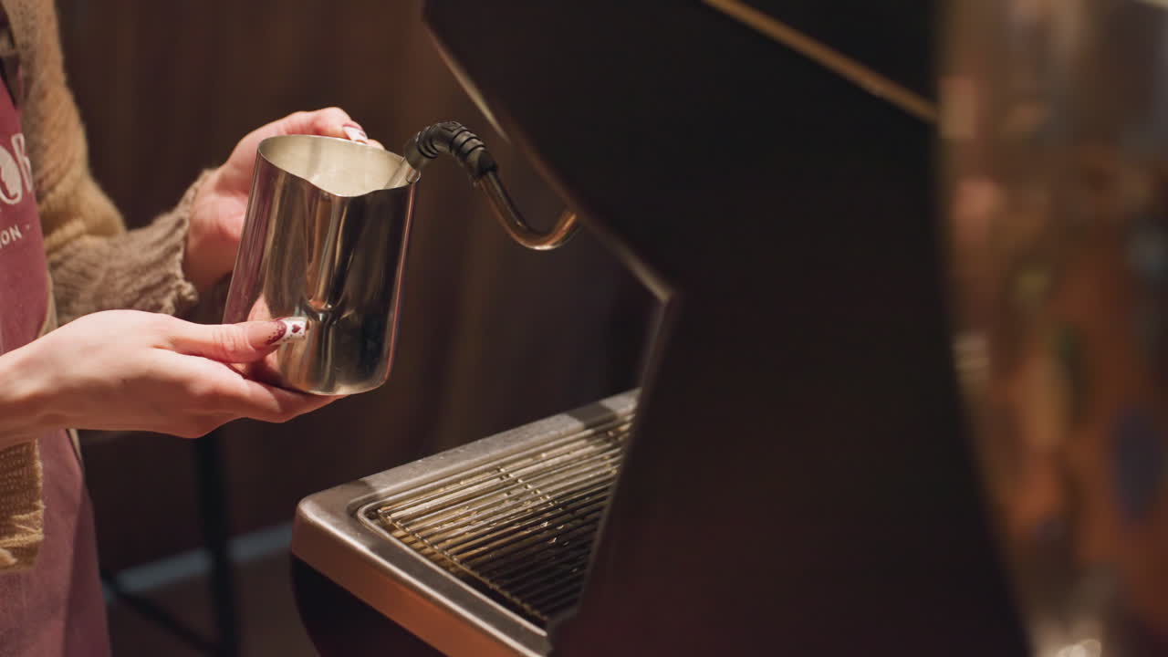 Close up of bar attender holding metal jug under electric water dispenser collecting hot water, visible water droplets falling as steam rises during beverage preparation routine