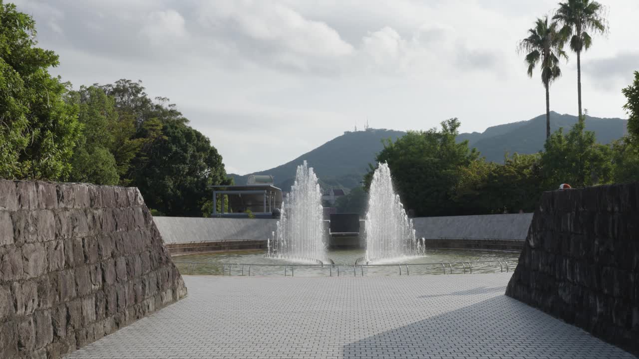 Joven caucásico caminando hacia una fuente en el Parque de la Paz de Nagasaki en un día soleado y mirando el monte Inasa, Japón