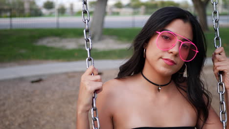 Eine-Schöne-Junge-Hispanische-Frau,-Die-Auf-Einem-Parkspielplatz-Spielt-Und-Schwingt,-Trägt-Eine-Rosafarbene-Retro-Sonnenbrille-In-Zeitlupe