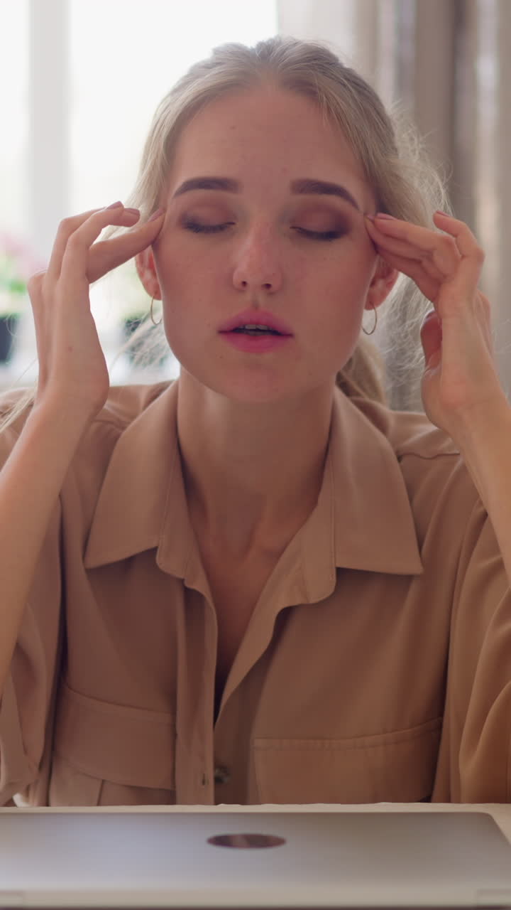Tired young woman having headache sits by closed laptop at white table massaging weary forehead with hands against window in room slow motion