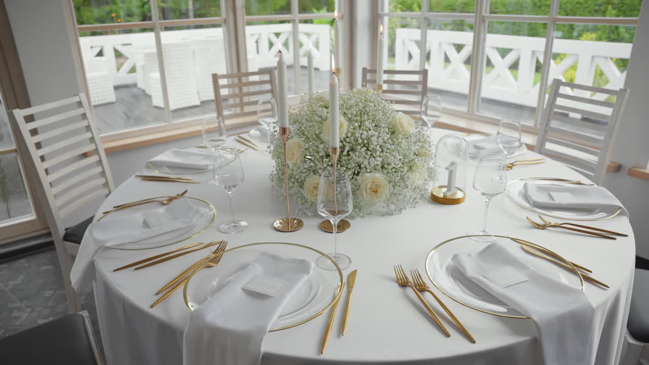 Elegant wedding reception table setup with white roses, baby's breath centerpiece, gold cutlery, and stylish glassware in a bright indoor venue with natural light and garden view
