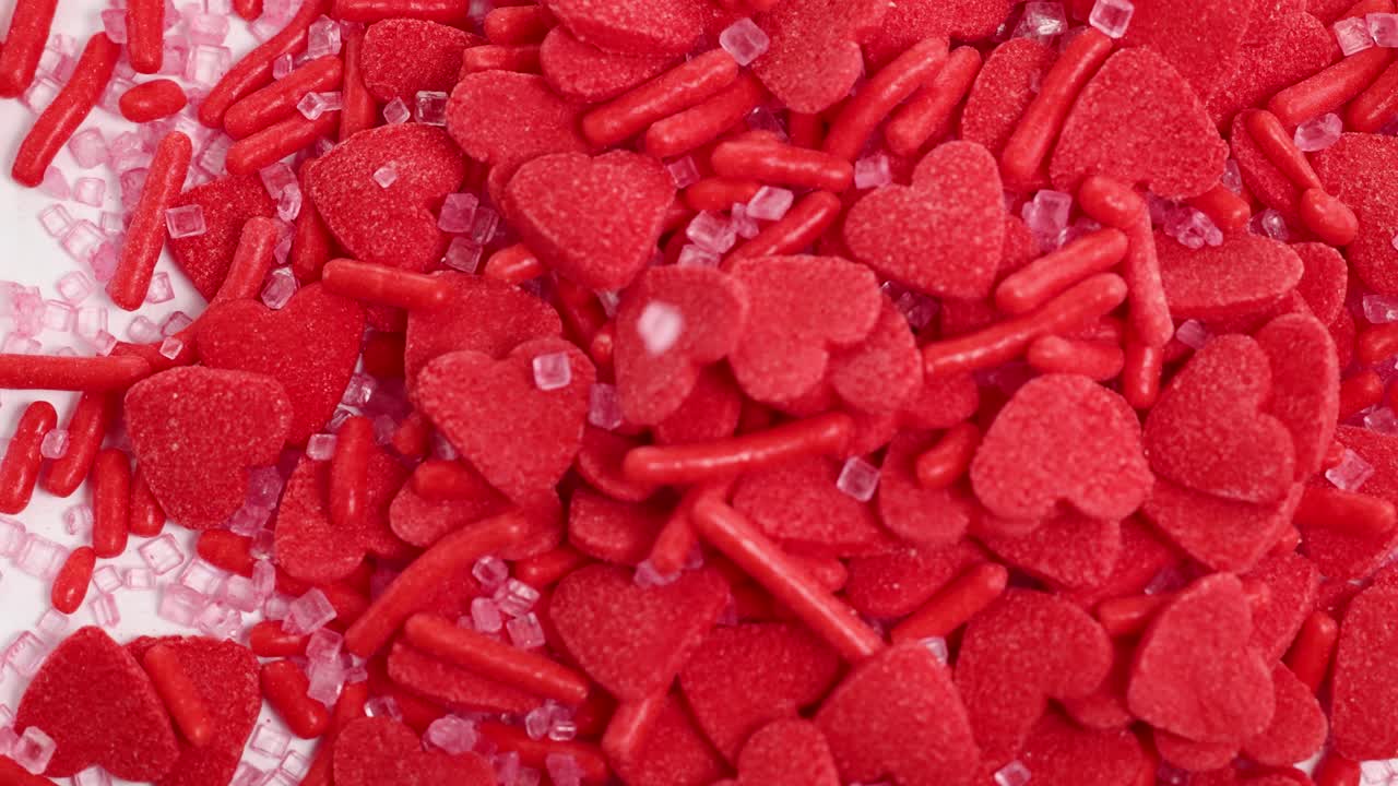 Red candy hearts and sprinkles cascade onto a white surface, creating a vibrant and playful visual effect